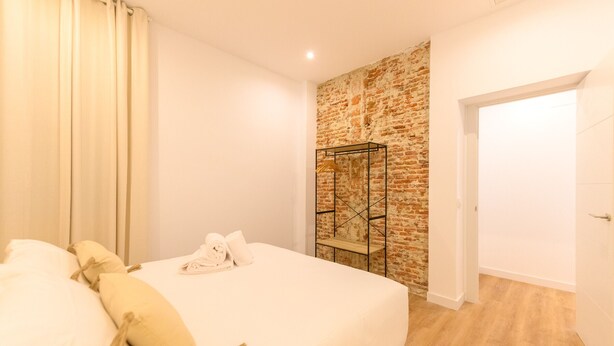 A bedroom with a brick wall, a metal rack with clothes, a bed with white linens, and a wooden floor.
