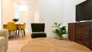 A modern living room with a round wooden coffee table, a green armchair, yellow chairs, a wooden cabinet, and a television.