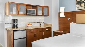 A compact hotel room with a kitchenette, including a microwave, coffee maker, and small refrigerator. There is a bed with a headboard, a bedside table with a lamp, and a painting on the wall.