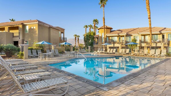 A pool area with lounge chairs and umbrellas, surrounded by buildings with balconies and palm trees.