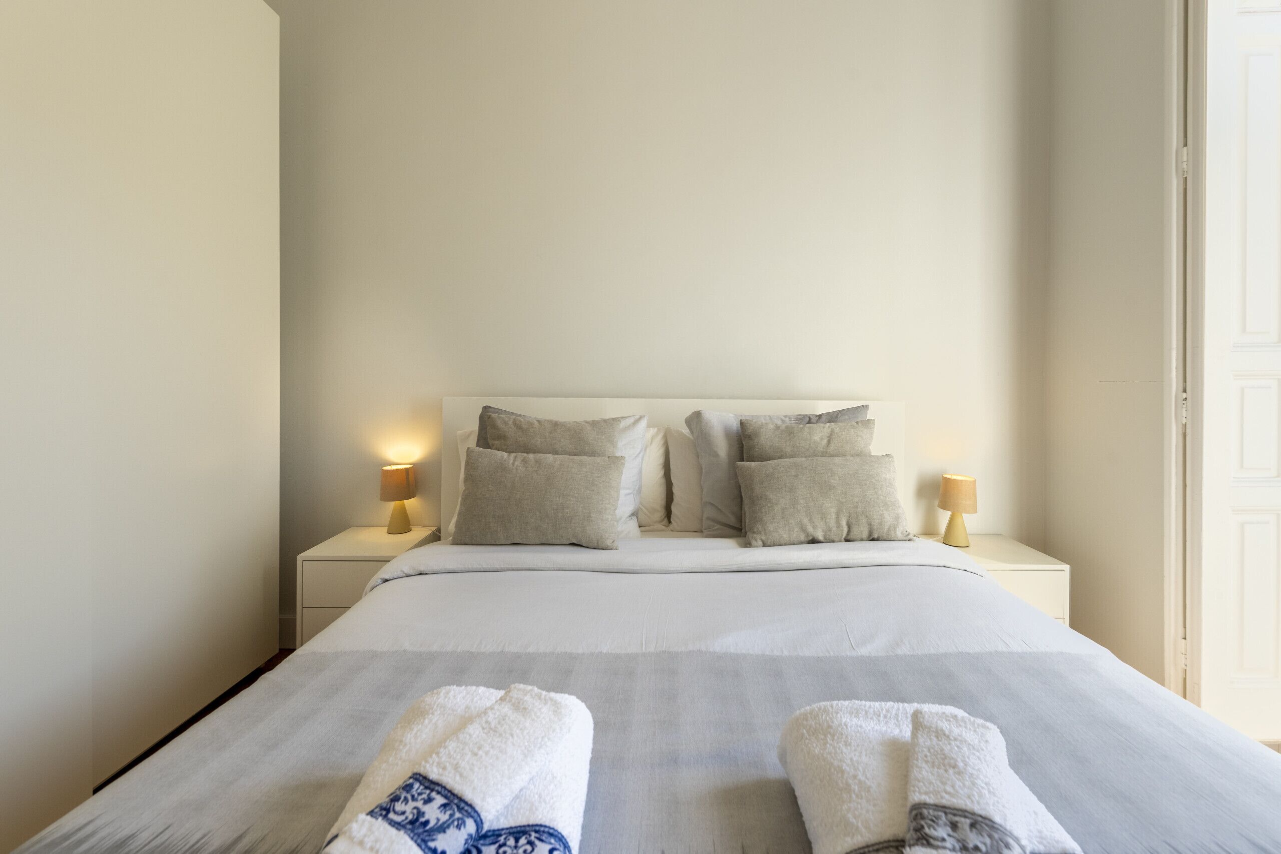 Una cama bien hecha con colcha a rayas blancas y grises, dos lámparas de noche y una mesita de noche blanca.