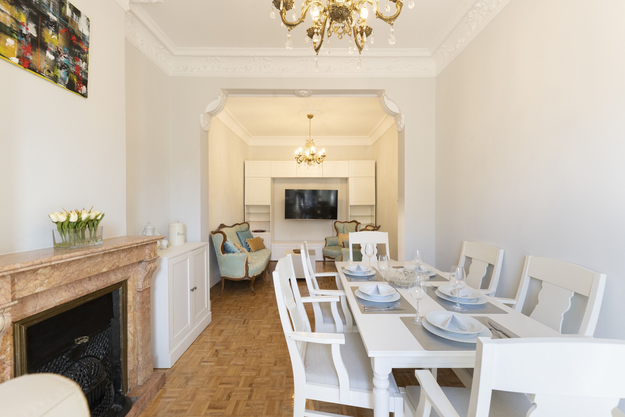 Un comedor con una mesa blanca dispuesta para seis personas, flanqueada por sillas blancas, una chimenea y un televisor al fondo.