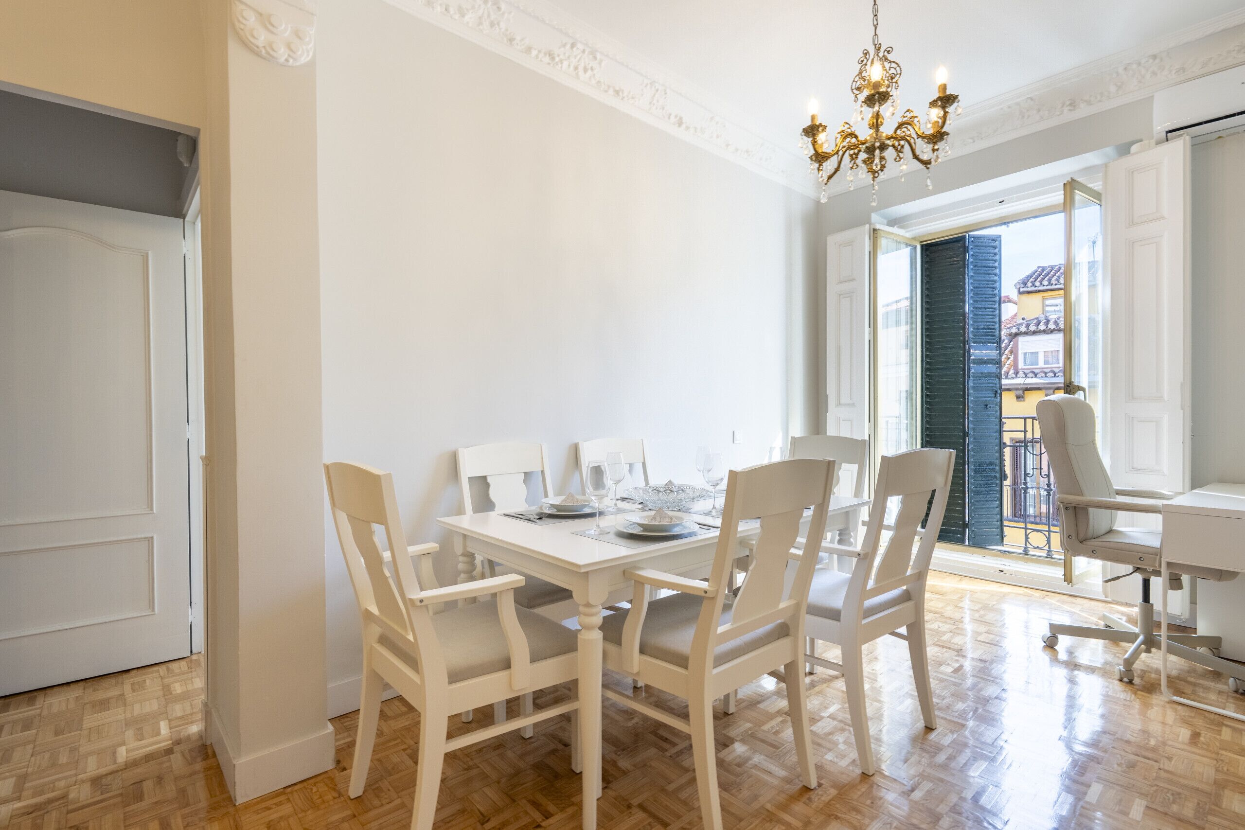 Un comedor con una mesa blanca dispuesta para seis personas, rodeada de sillas blancas. Encima de la mesa hay una araña y se ve el exterior a través de un puerta abierta.