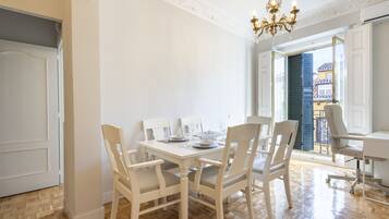 Un comedor con una mesa blanca dispuesta para seis personas, rodeada de sillas blancas. Encima de la mesa hay una araña y se ve el exterior a través de un puerta abierta.
