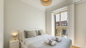 En pænt redt seng med puder og et tæppe, et sengebord med en lampe, og udsigt til bygninger gennem et vindue.