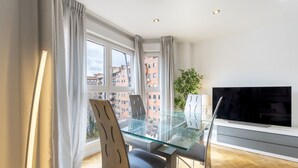 A modern living room with a glass dining table, two chairs, a flat-screen TV, and a large window with curtains.