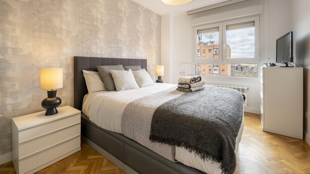 A modern bedroom with a large bed, bedside lamps, a radiator, a television, and a view of buildings through a window.