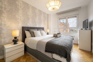 A modern bedroom with a large bed, bedside lamps, a radiator, a television, and a view of buildings through a window. - Spacious 3-bedroom, 2-bathroom flat in Residential Complex with Swimming Pool, Paddle Tennis _ Madrid  (Madrid)