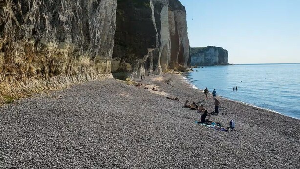 A pebbled beach with people sunbathing and swimming, bordered by tall cliffs.