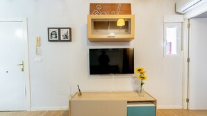 A room with a wooden TV stand, a mounted TV, a small shelf with a lamp, and a vase with sunflowers.