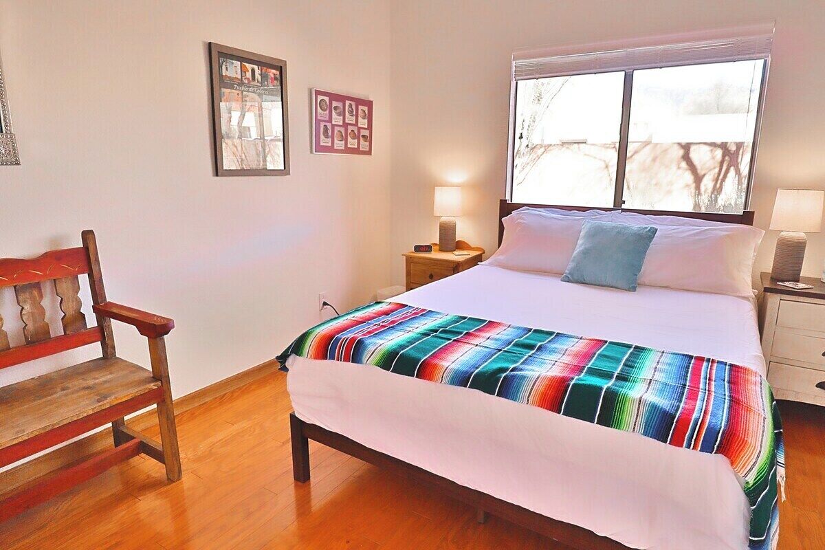 A bedroom with a wooden bed, a colorful striped blanket, a wooden chair, a nightstand with a lamp, and a window with a view.