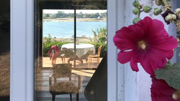 Una porta vetrata conduce a una zona con posti a sedere all'aperto, con tavolo e sedie, con vista su una spiaggia e il mare.
