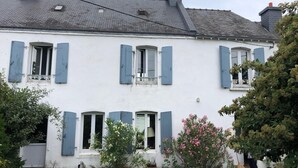 Un edificio bianco a due piani con persiane blu, tetto in tegole e un giardino con diverse piante e fiori.