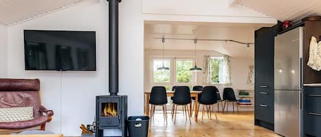 A modern living room with a wood-burning stove, a dining area with chairs, a TV mounted on the wall, and a kitchen with a refrigerator.