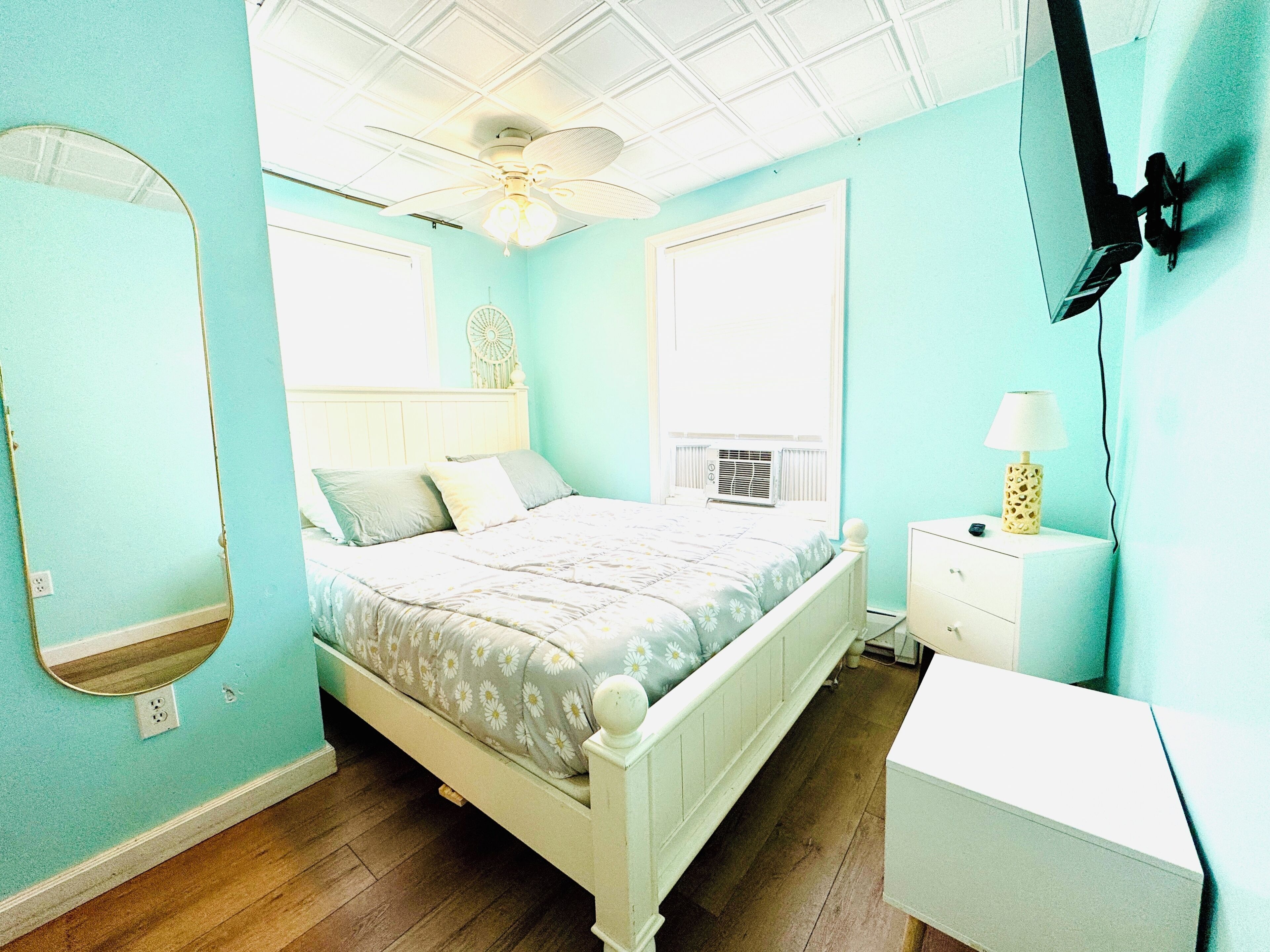 A bedroom with a bed, a nightstand, a mirror, a ceiling fan, and a wall-mounted TV.