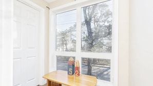 Una habitación con un banco de madera y una puerta, una ventana con vistas a árboles y una carretera, y una puerta blanca.
