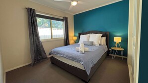 Une chambre à coucher comprenant un lit, un ventilateur de plafond, une fenêtre avec des rideaux, une lampe de chevet et une petite table avec une lampe.