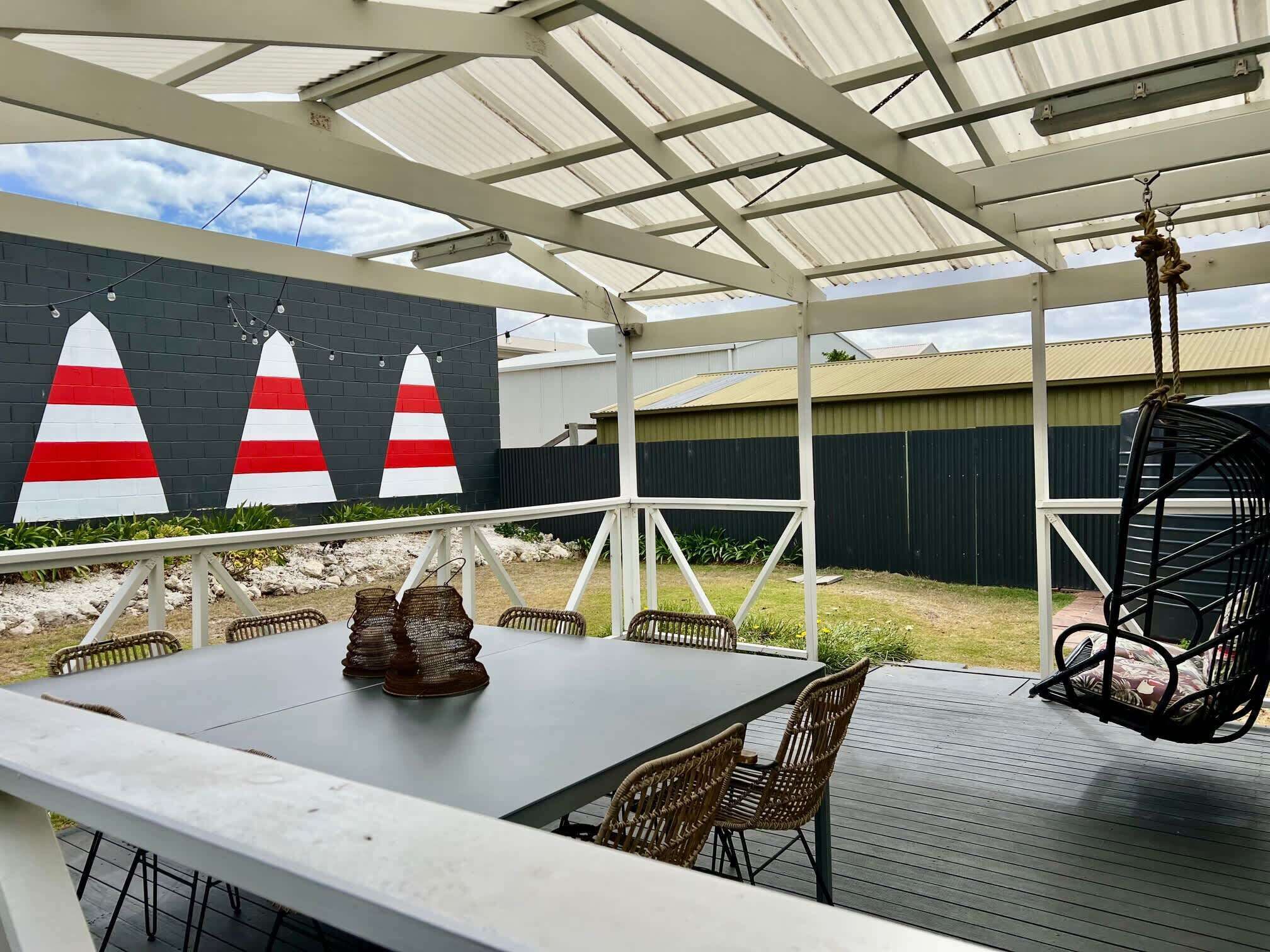 Une terrasse extérieure avec une table et des chaises, un espace abrité avec un toit en métal, et une fresque représentant des voiliers sur un mur.