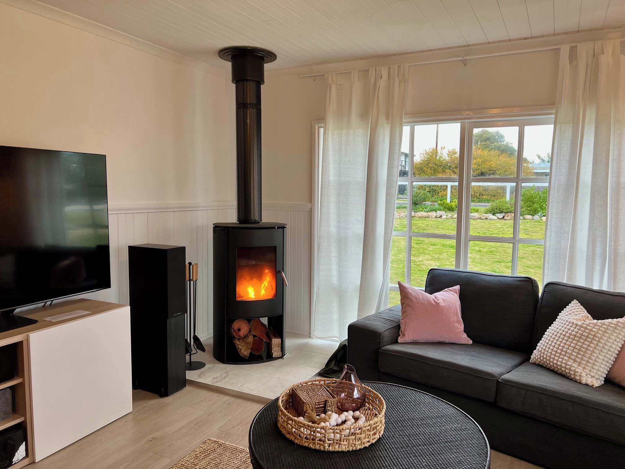 A living room with a black wood-burning stove, a flat-screen TV, a grey sofa, and a large window with curtains.