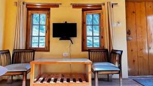 Een kamer met houten plafond en wanden, een aan de muur bevestigde televisie, een houten tafel met stoelen en ramen voorzien van gordijnen.