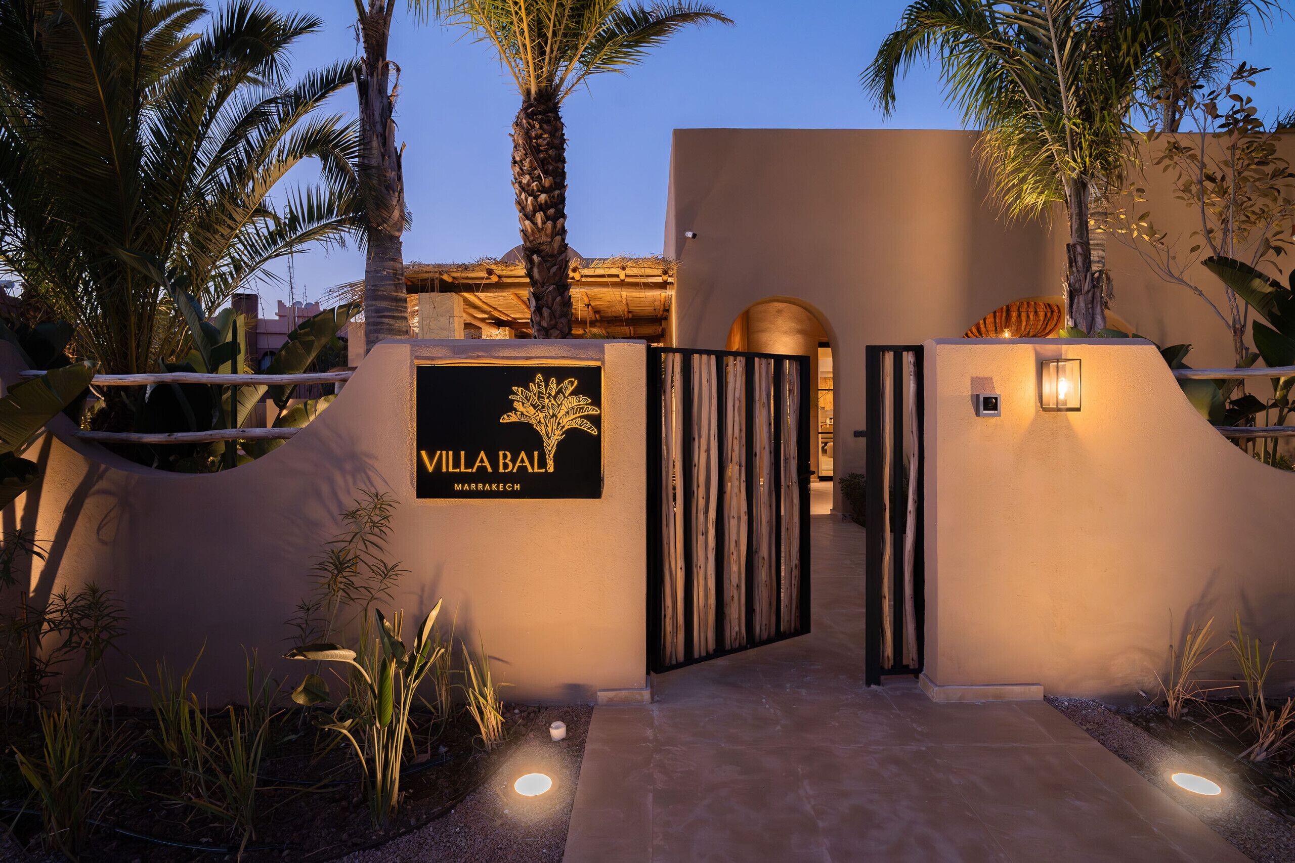 A gated entrance to Villa Bal in Marrakech with a sign, palm trees, and a well-lit pathway.