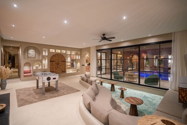 A modern living room with a foosball table, comfortable seating, and a view of an indoor pool. - Villa BALI with private heated pool, sauna, close to Amelkis golf course (Marrakech)