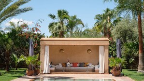 Un espace piscine doté d’un coin salon abrité, entouré de palmiers et de plantes en pot.