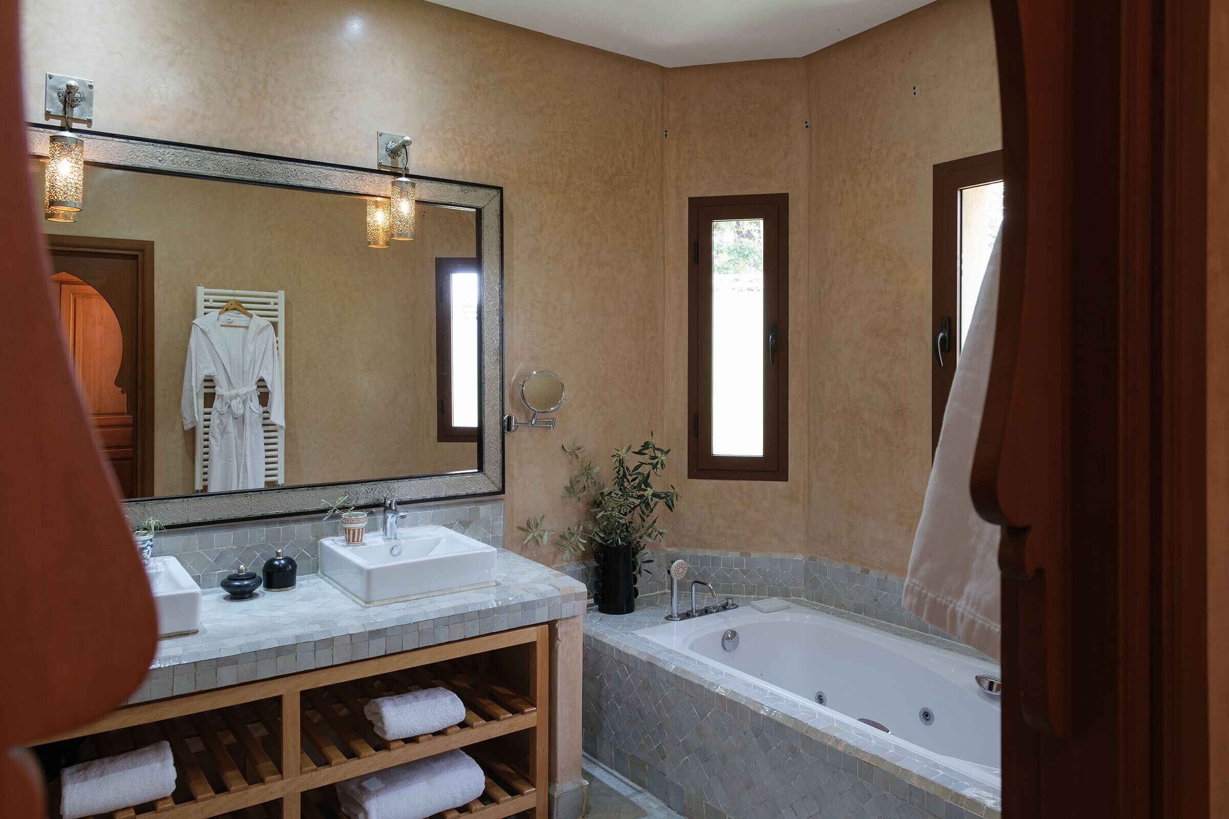 Une salle de bain avec deux lavabos, un grand miroir, une baignoire et une étagère en bois où sont posées des serviettes.