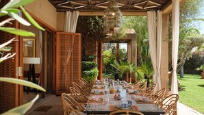 Un espace de restauration en plein air avec une longue table dressée pour accueillir plusieurs convives, entourée de chaises en osier, sous une pergola agrémentée d’un luminaire suspendu.
