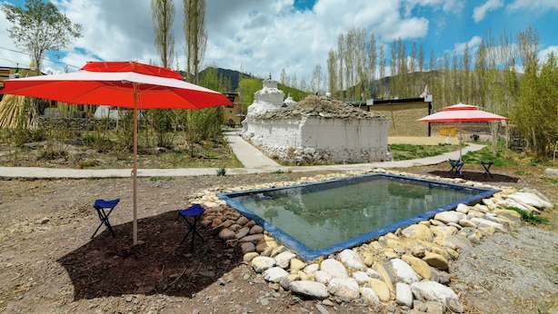 A swimming pool surrounded by rocks, with red umbrellas and blue chairs. - House Of Stok (Leh)
