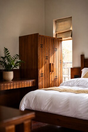 Un dormitorio con un armario de madera, una mesita de noche con una planta, una cama con ropa de cama blanca y una ventana con persiana.