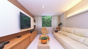 A modern living room with a flat-screen TV, a beige sofa, a wooden coffee table with a plant, and a window with curtains. - Aristides Leblon (Rio de Janeiro)