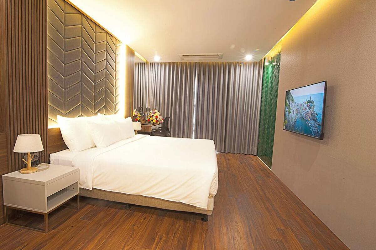 A modern hotel room with a large bed, a nightstand with a lamp, a wall-mounted TV, and a view of the sea through the curtains.