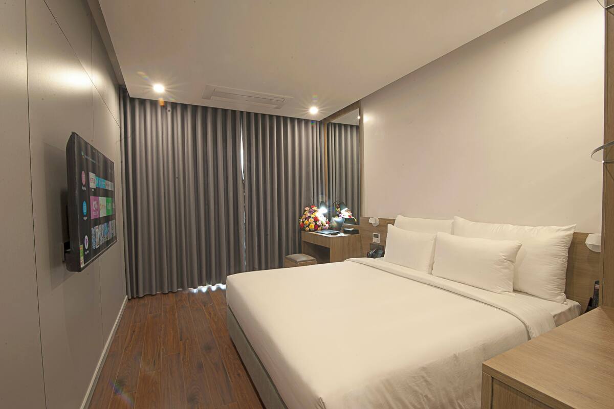 A modern hotel room with a large bed, a desk with a lamp, a mirror, and a wall-mounted TV.