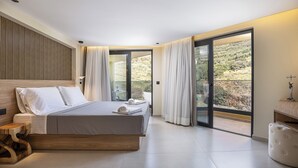 A modern bedroom with a large bed, a wooden headboard, a bedside table, and a view of a natural landscape through large sliding glass doors. - Elmima Villa  (Mylopotamos)