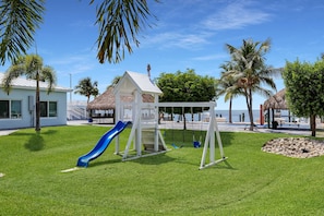 A playground with a slide and swings, surrounded by palm trees and a building with a thatched roof.