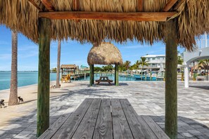 A wooden deck with a thatched gazebo, a pool area with boats, and a beachside view.