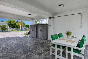 An outdoor patio with a white table and green chairs, a grey storage unit, and a clear view of the surrounding area.