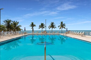 A swimming pool with lounge chairs and palm trees, overlooking a clear blue sky and ocean.
