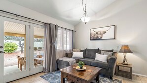 A living room with a sofa, coffee table, and a lamp. There is a sliding glass door leading to an outdoor area with a patio table and chairs.