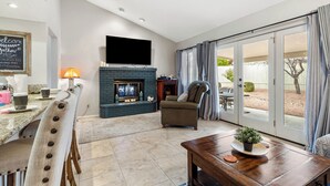 A living room with a fireplace, a television mounted on the wall, a dining area with a table and chairs, and a sliding glass door leading to an outdoor area.