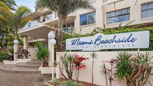 A multi-story building with a sign for Miami Beachside Holiday Apartments. There are steps leading to the entrance, surrounded by various plants and a palm tree.