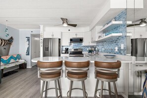 Uma cozinha moderna com armários brancos, eletrodomésticos de aço inoxidável, uma ilha central com banquetas e um ventilador de teto.