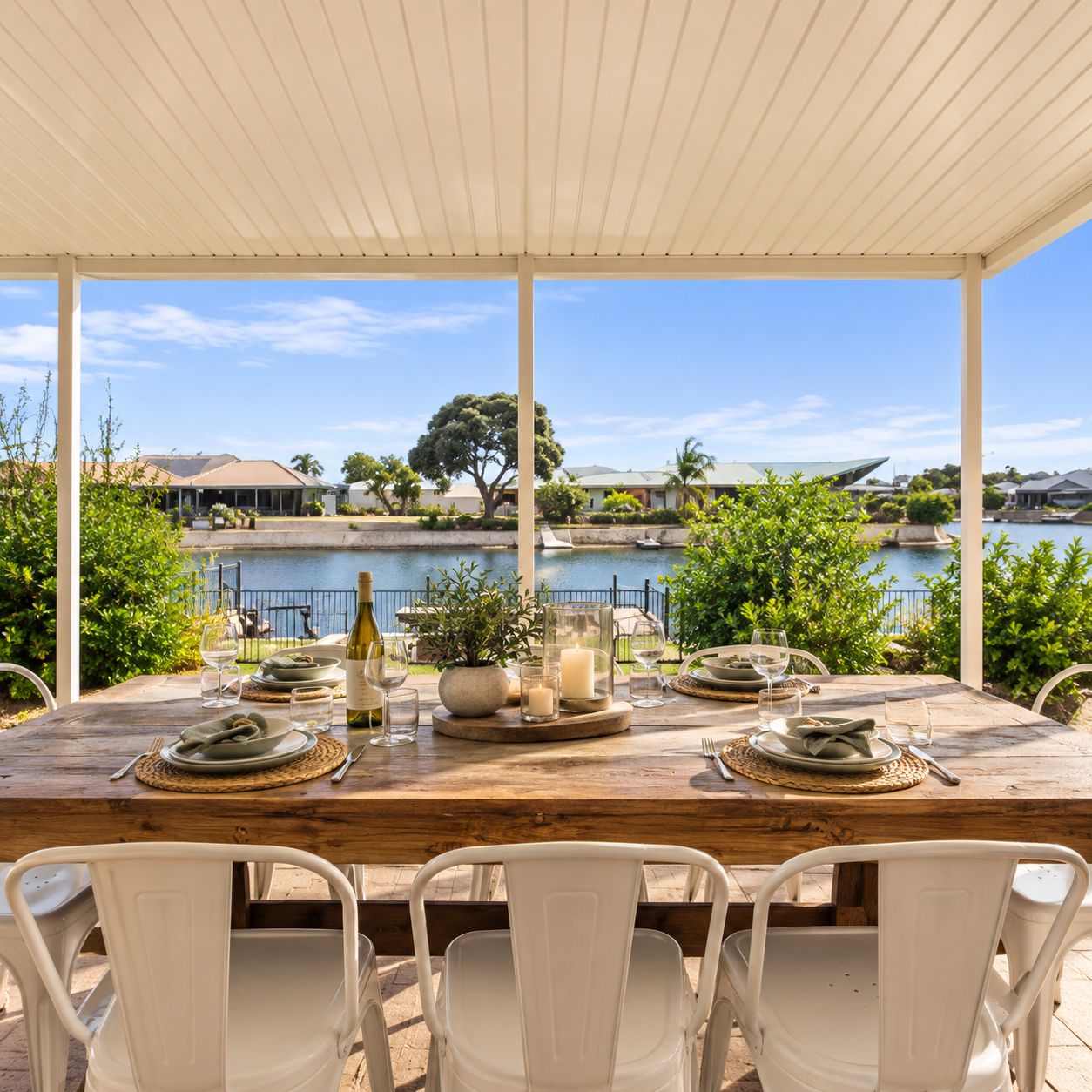 An outdoor dining area with a view of a lake, a wooden table set with plates, glasses, and a bottle, surrounded by white chairs.