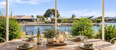 An outdoor dining area with a view of a lake, a wooden table set with plates, glasses, and a bottle, surrounded by white chairs.
