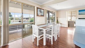 A spacious room with a large sliding glass door leading to a balcony, a dining area with white furniture, and a view of a water body and greenery outside. - Canal view • Pet friendly • Private Jetty • Spacious Yard (South Yunderup)