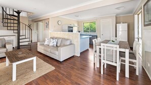 A spacious living area with a sofa, coffee table, and dining table. A kitchen with a refrigerator and stove is visible in the background. - Canal view • Pet friendly • Private Jetty • Spacious Yard (South Yunderup)