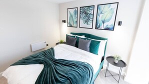 Une chambre moderne avec une tête de lit verte, un couvre-lit blanc et trois reproductions botaniques encadrées accrochées au mur.