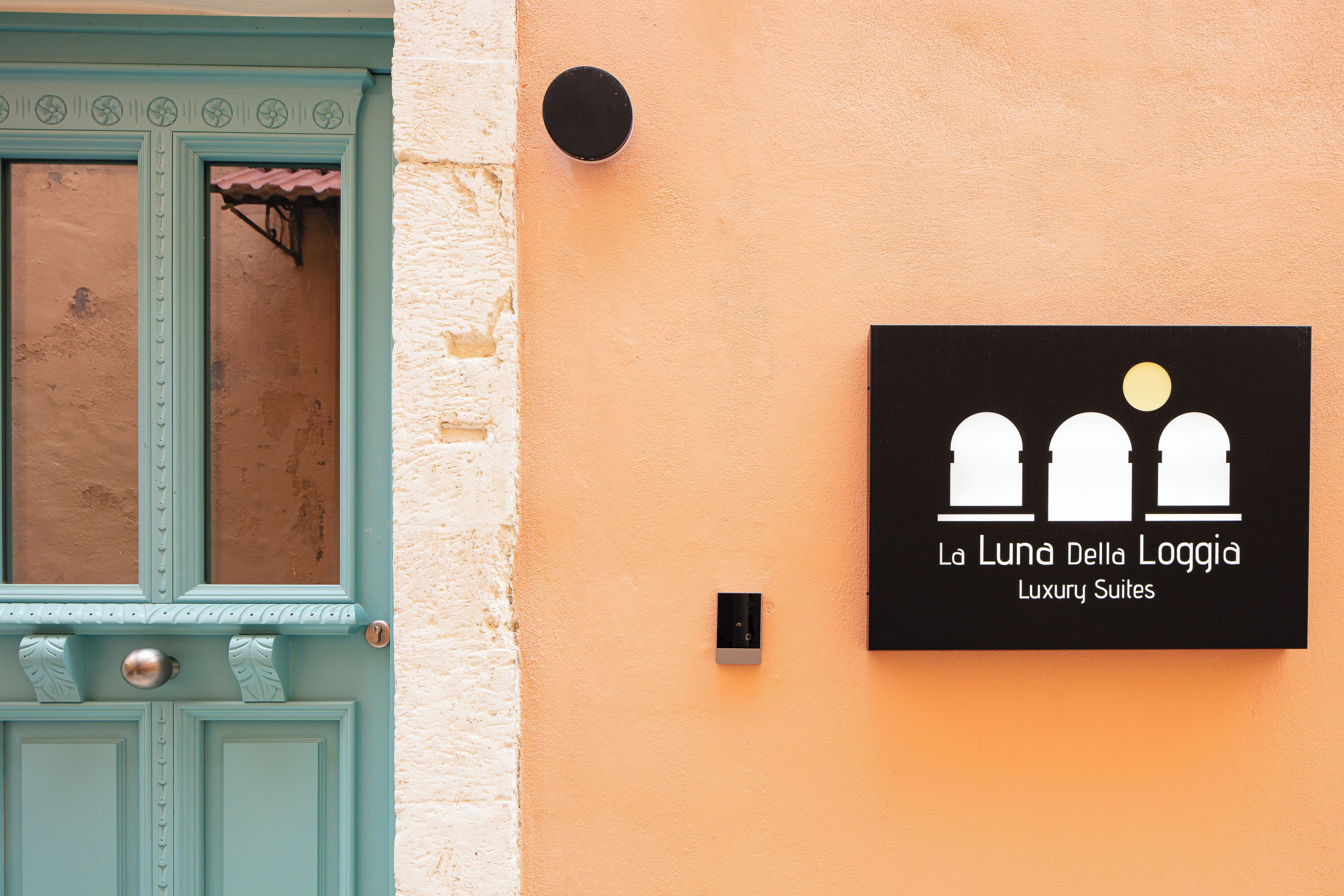 Une porte de couleur bleu sarcelle avec une fenêtre, une enseigne noire avec du texte blanc et un luminaire fixé au mur.
