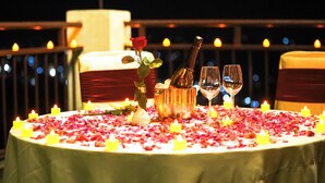 A table set for two with candles, a bottle, and glasses, surrounded by rose petals. - Hotel Dhroov By Mystical Views (Shimla)